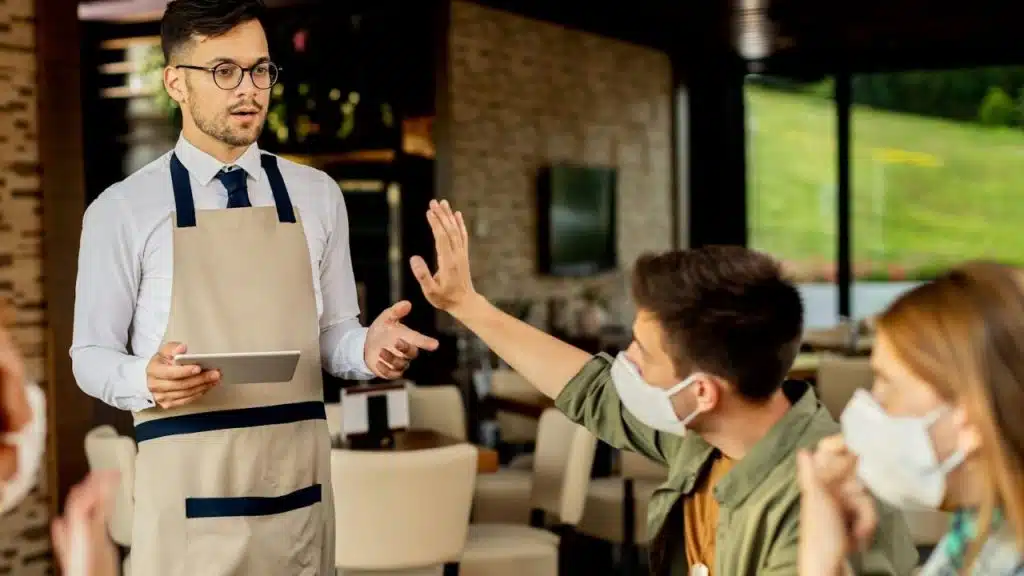Man in a face mask gesturing stop to a waiter holding a tablet in a restaurant.