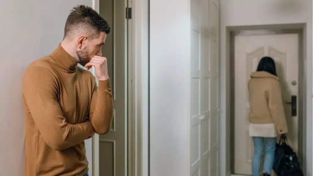 A man looking sad or worried as a woman walks away and out the front door, carrying a bag.