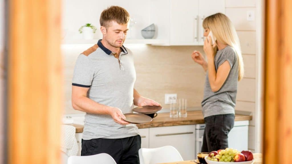A man holds two plates in a kitchen while a woman stands behind him talking on a phone.