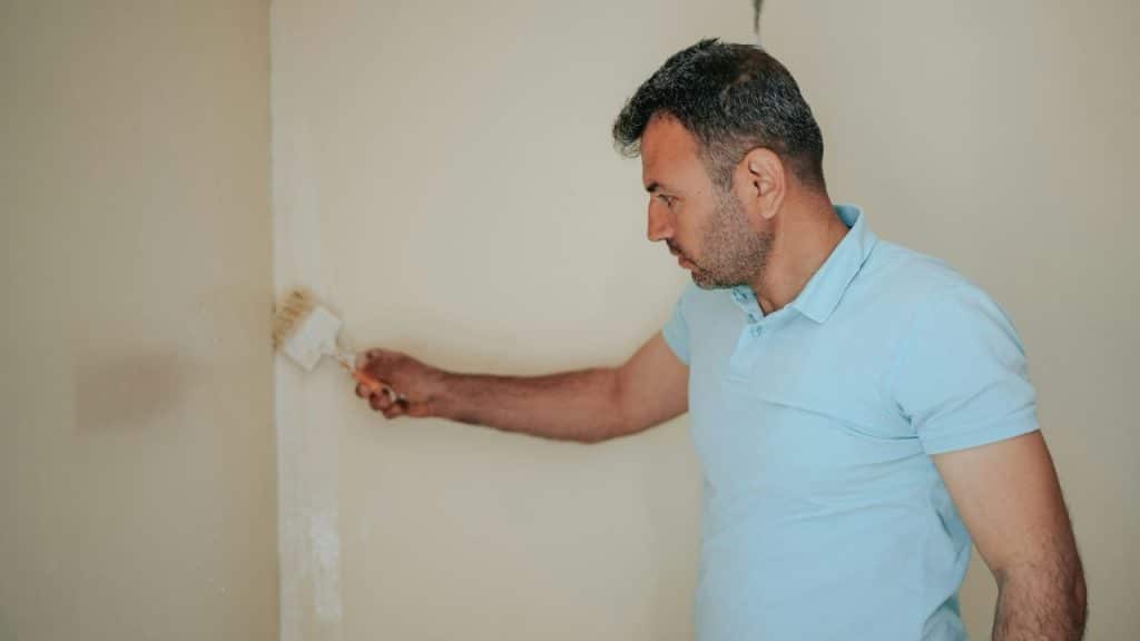 Man in a light blue polo shirt applies white paint to a wall with a brush.
