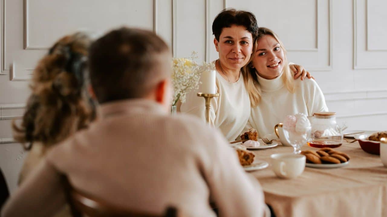 Two awkward women embrace at a dining table with others blurred in the foreground.