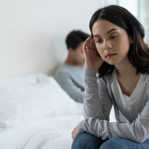 A woman looking sad and troubled while sitting on a bed with a man turned away in the blurry background.