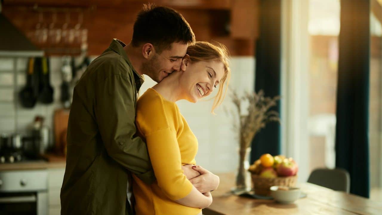 Man embracing a smiling woman from behind in a sunny, rustic kitchen setting.