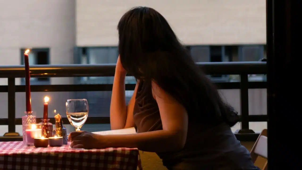 Woman with long dark hair sitting alone at a candlelit table on a balcony with a glass of wine.