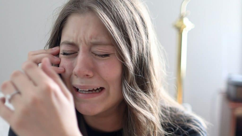 A crying woman with long hair holds her hand to her head, with a gold lamp visible in the background.
