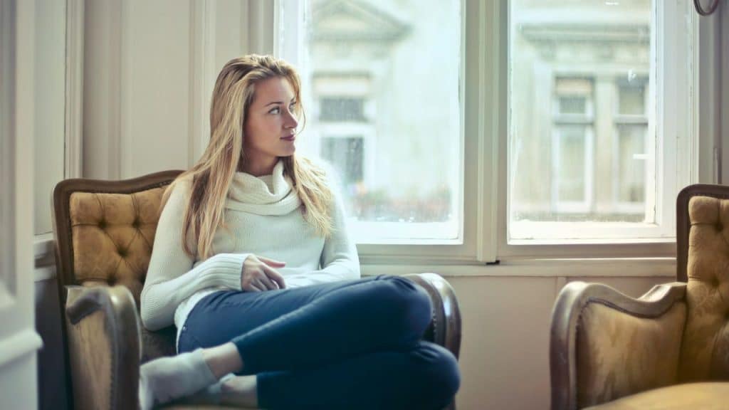 Woman in a white turtleneck and blue jeans sits in a chair looking out a window.