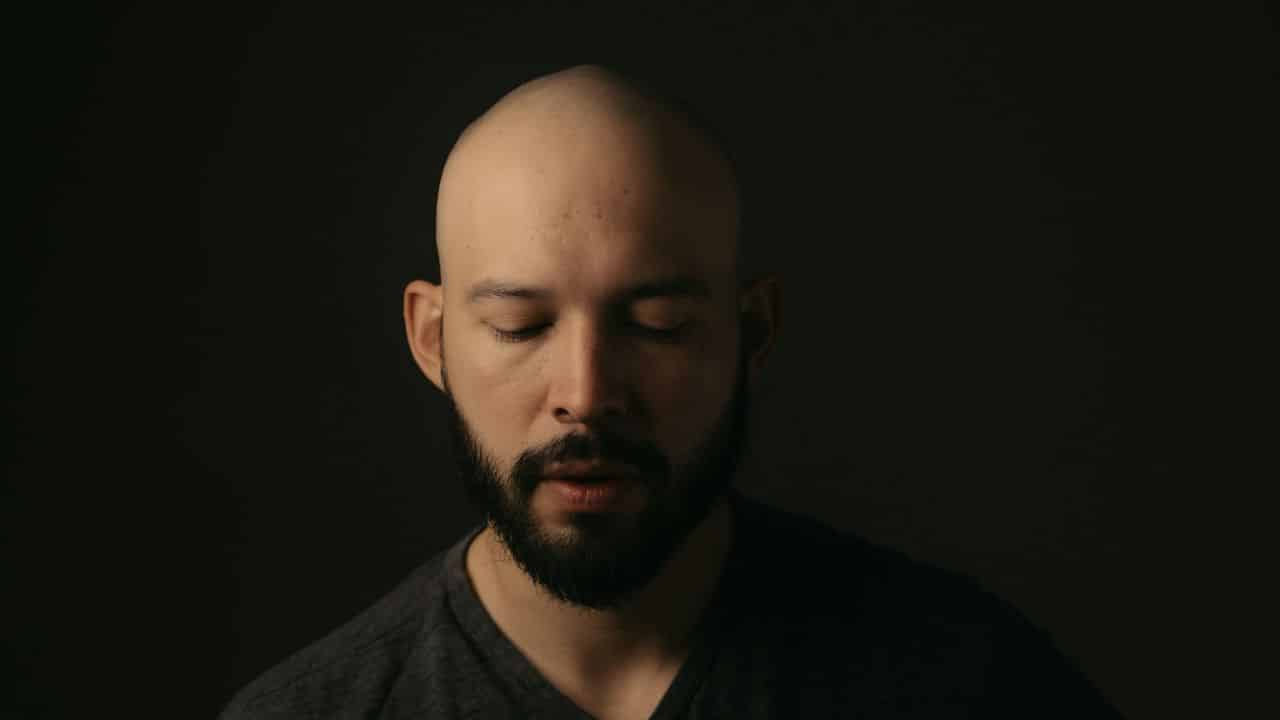 Close-up of a bald man with a beard and closed eyes on a dark background.