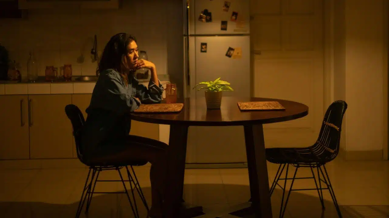 A woman sits alone at a dark kitchen table, resting her chin on her hand, looking distressed.