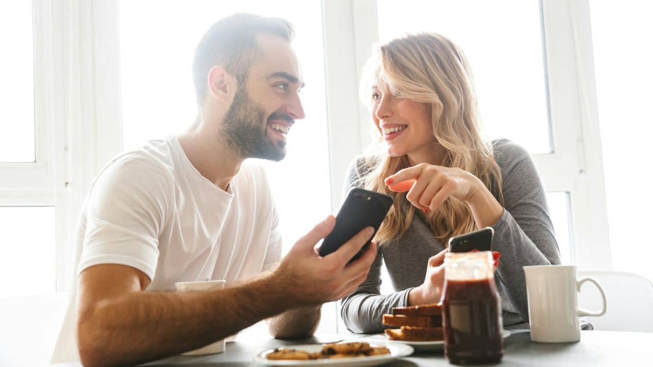 A smiling man and woman are sitting at a table looking at their phones during a meal.