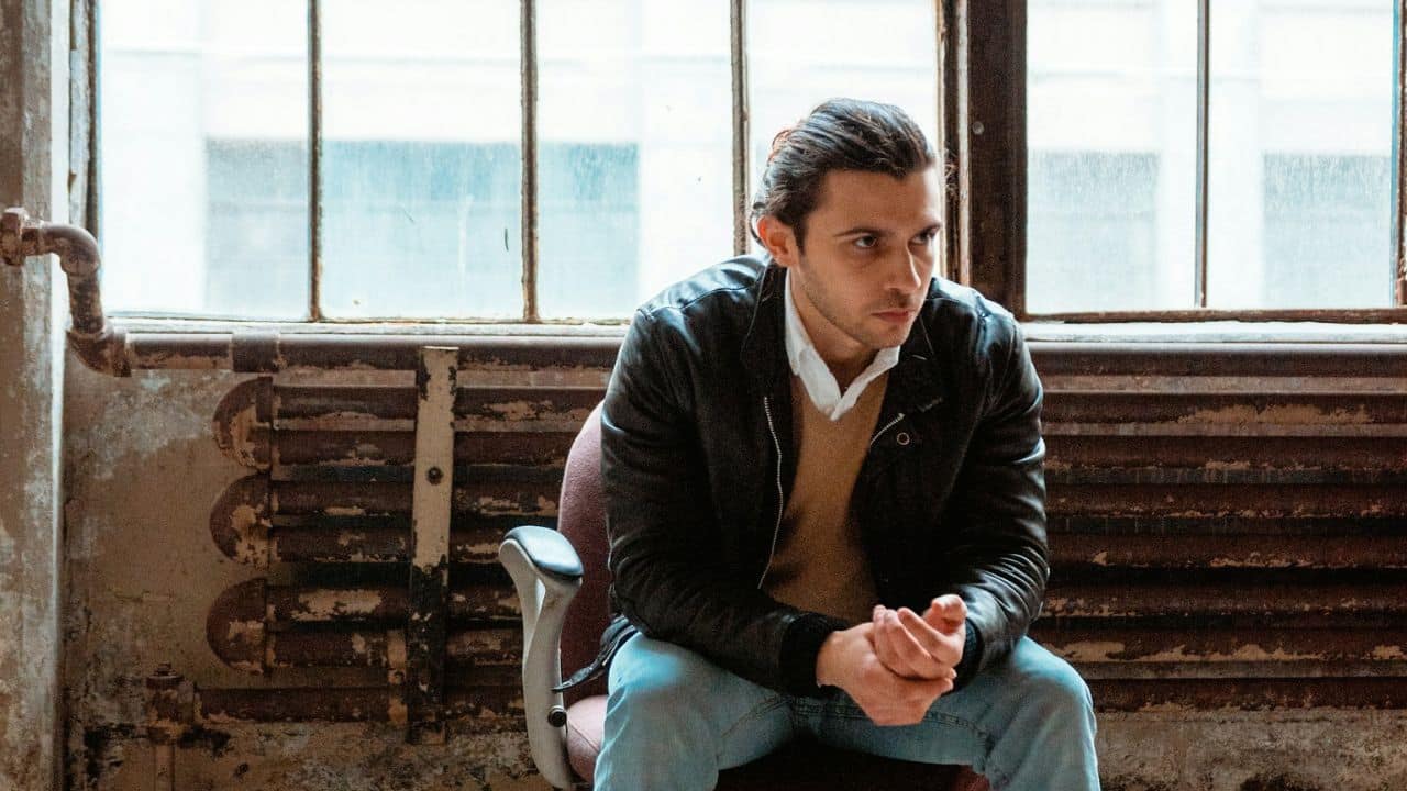 A man sits in a chair in front of a window in an industrial-looking room.