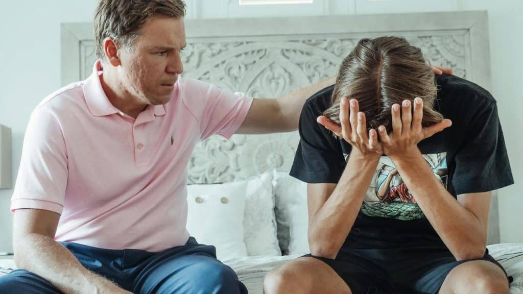A father comforting his son, who is sitting with their face in their hands.