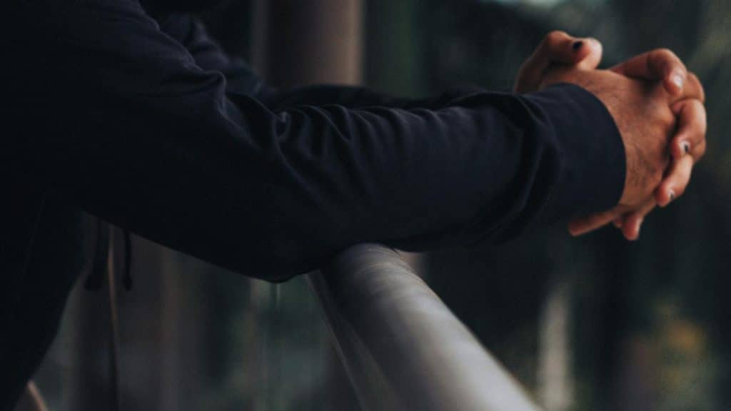 A person leans on a railing with hands clasped, wearing a dark long-sleeve shirt.