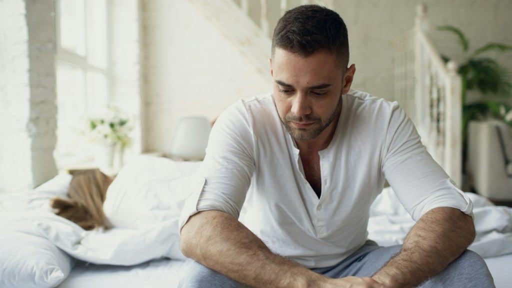 A man sitting on a bed looking down thoughtfully.