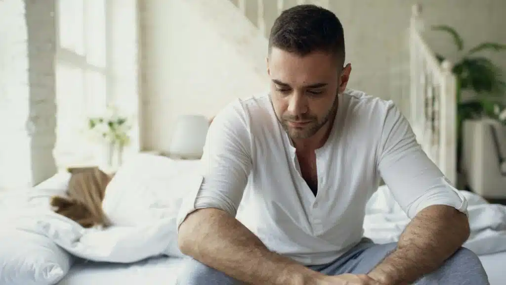 A distressed man is sitting at the edge of the bed while his wife is sleeping in the background.