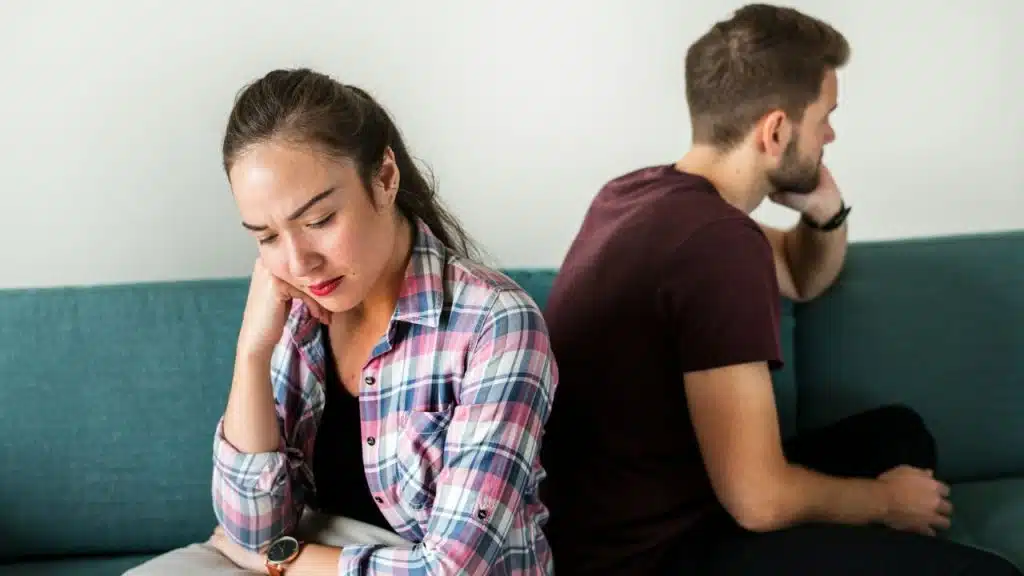A woman and man sit back-to-back on a teal couch, looking upset or distant.