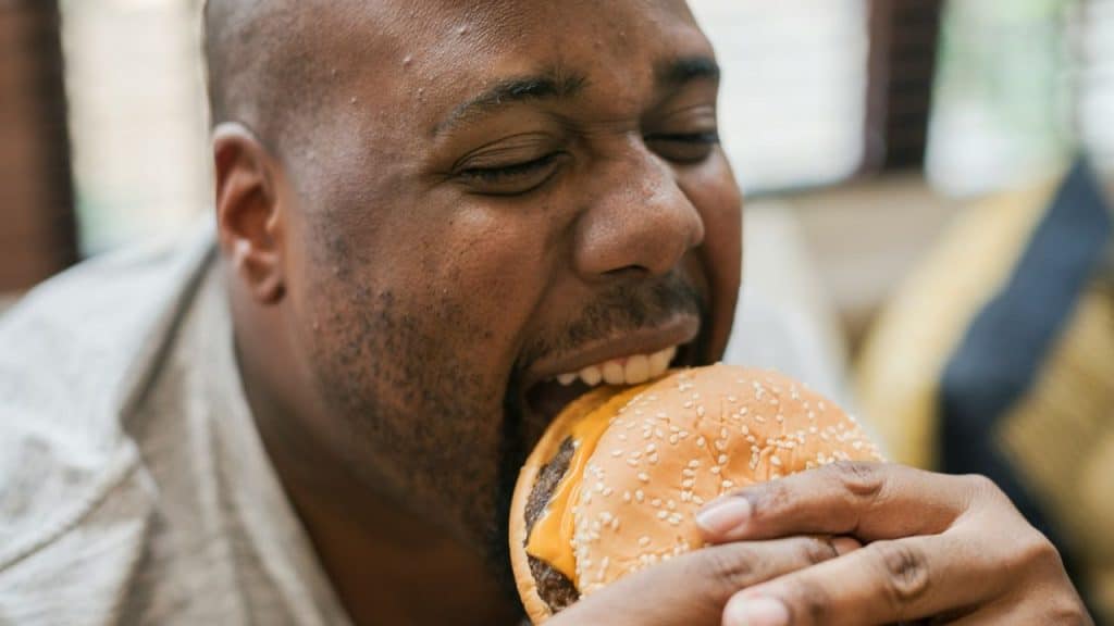 Close-up of a Black man with eyes closed biting into a cheeseburger.