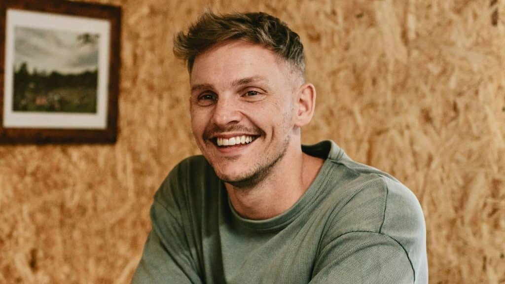 Young man smiling widely at the camera in a casual indoor setting with wood walls.