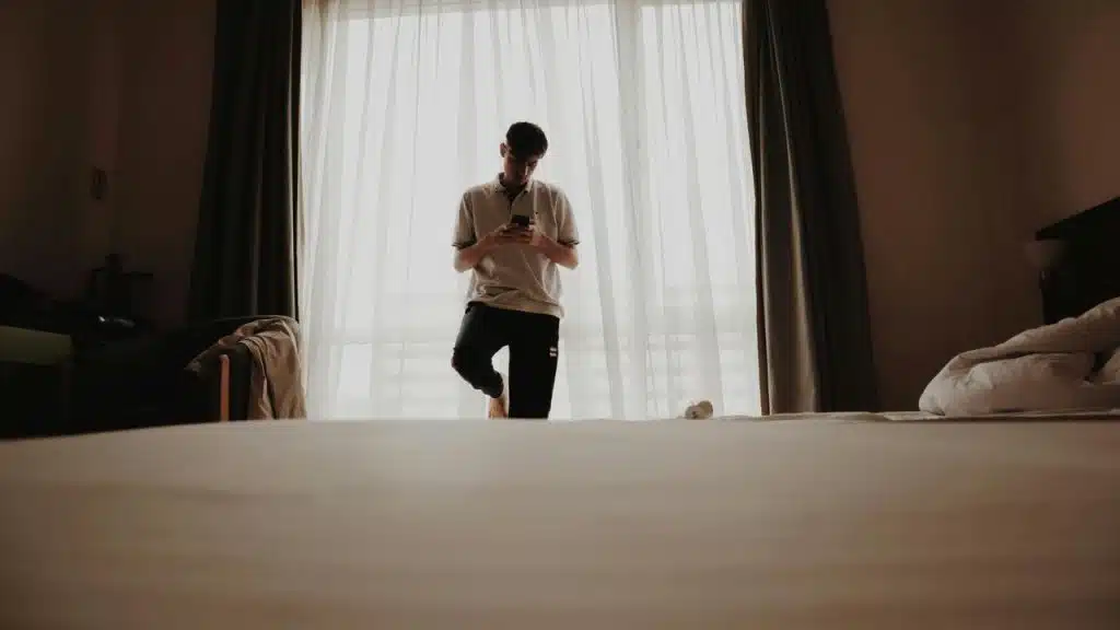 A young man standing by a window in a dim hotel room looking at his phone.