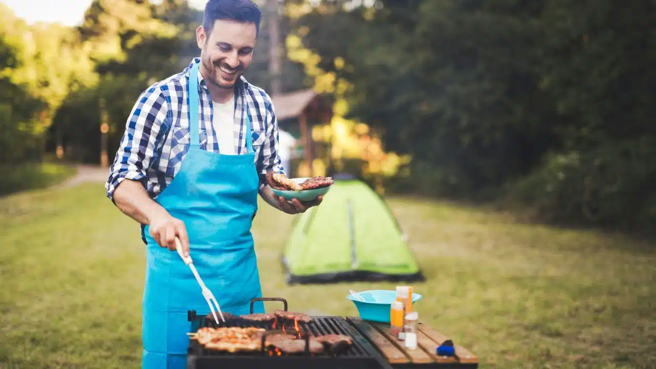 A smiling man in a blue apron grilling meat outdoors near a green tent.