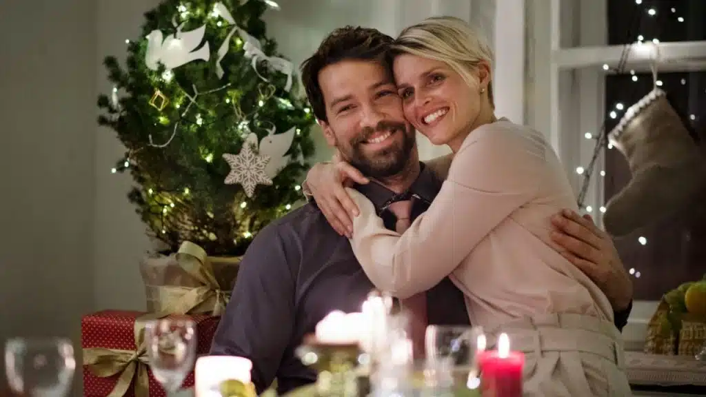 A smiling couple hugging at a candlelit dinner table with a decorated Christmas tree.