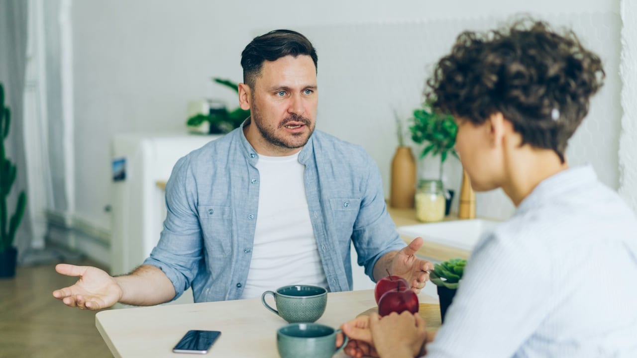 A man gestures with open hands while talking to a woman across a table with coffee cups.