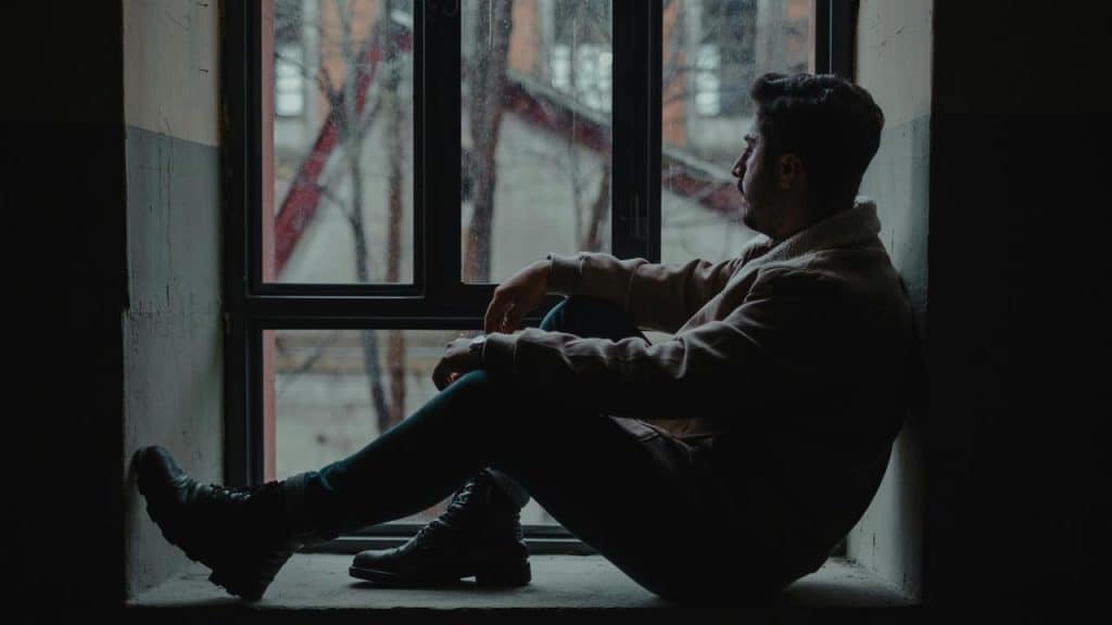 A man sitting in a dark windowsill, gazing outside thoughtfully.