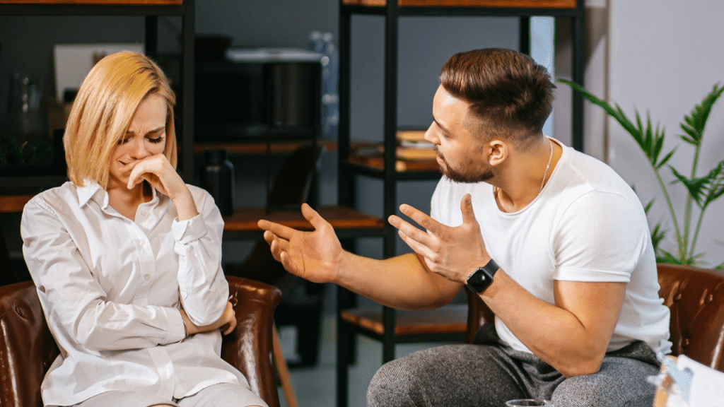 Woman sits upset, covering her mouth, while a man gestures and speaks toward her.