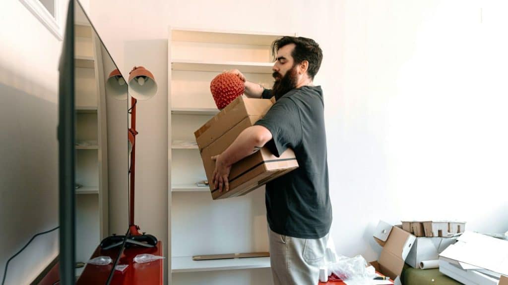 A man with a beard is carrying boxes and a red object near a white shelf.