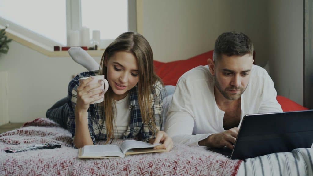 A couple is lying on a bed; the woman is drinking from a mug and reading a book, while the man is working on a laptop.