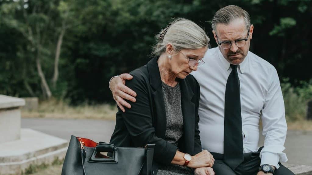 Middle-aged man in suit comforts older woman in black at an outdoor gathering.