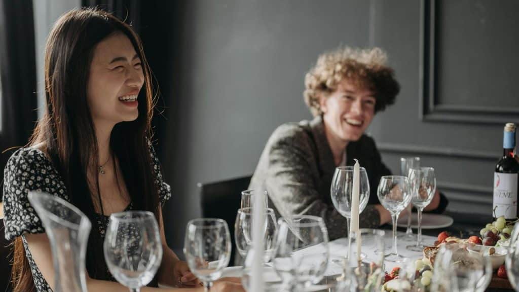 A photo of two people laughing together at a dinner table set with wine glasses and food..