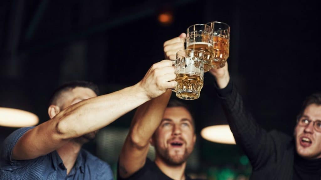 Three men in a dimly lit setting cheer, raising mugs of beer above their heads.