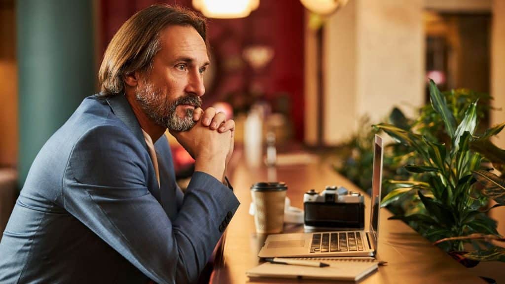 A middle-aged man in a blue suit jacket sits at a counter, looking intently off-camera.