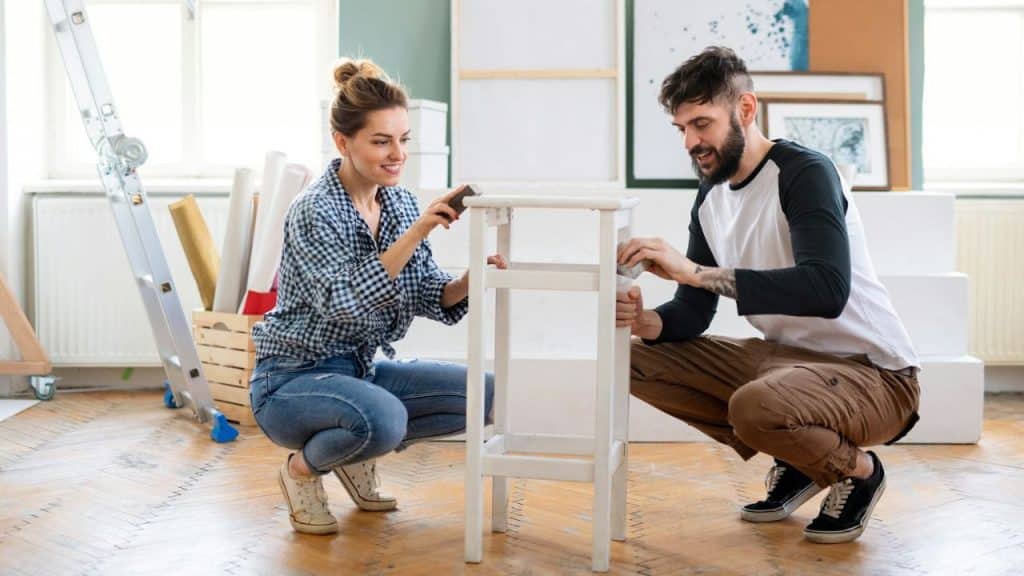 Smiling couple kneeling on a wooden floor, sanding and painting a small white table.