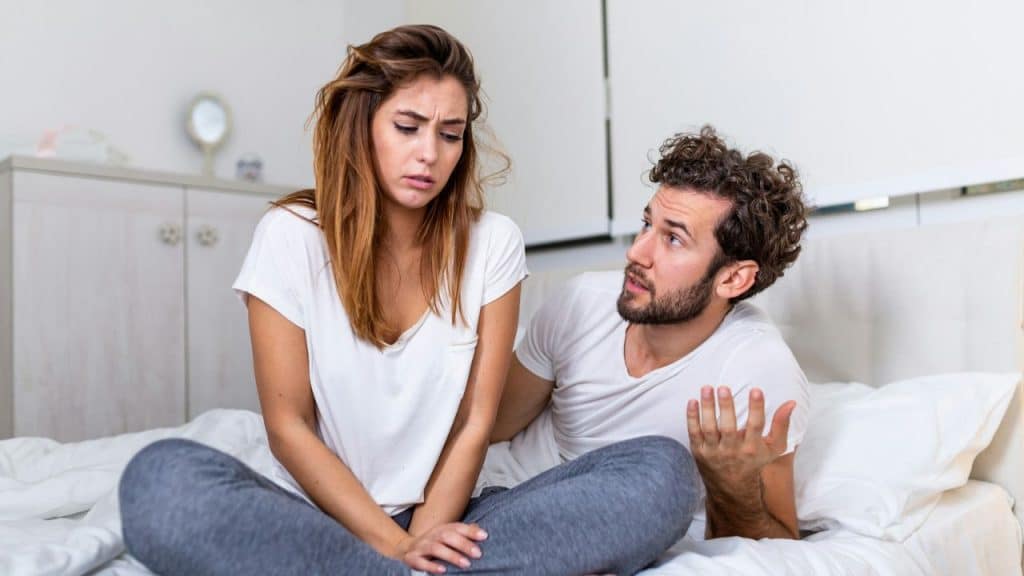 Woman in white shirt and grey pants looking distressed while man talks to her in bed.