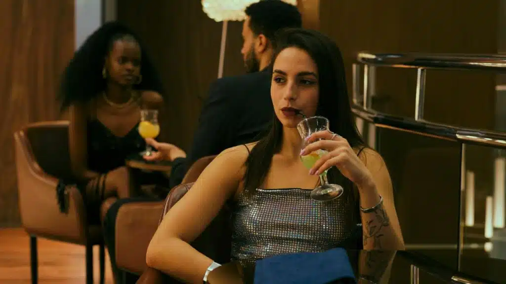 A woman in a sparkly top sips a drink in a dimly lit, upscale bar setting.