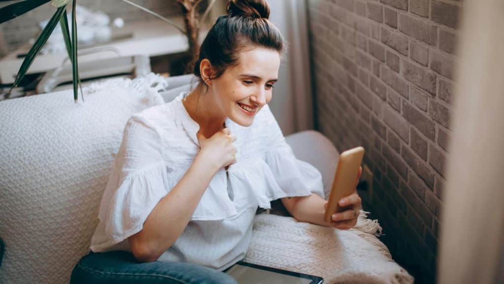 A woman smiling while holding a smartphone on a couch.