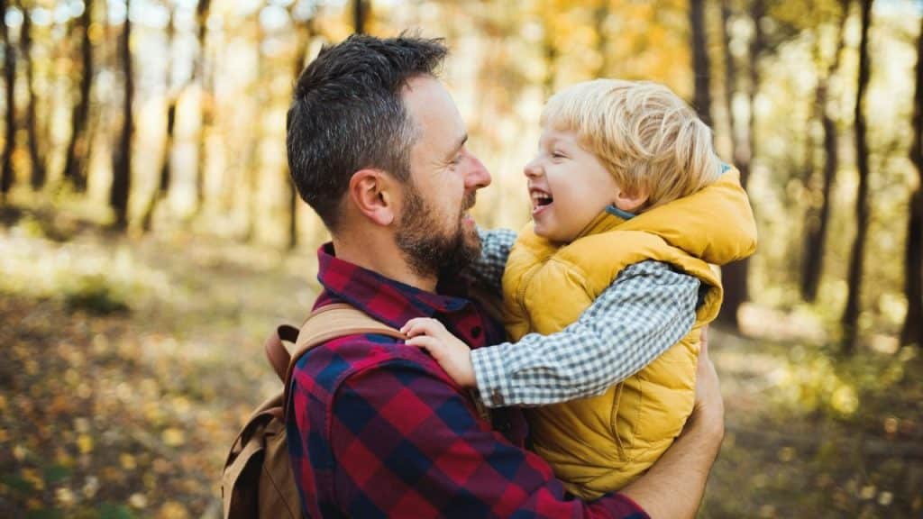A bearded father holds his laughing young son in a yellow vest in an autumn forest.
