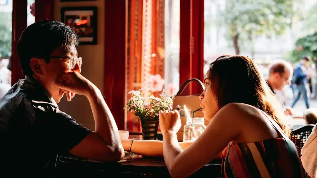 A man wearing a black-collared top sitting on a chair in front of a table and a woman wearing a multicolored top.