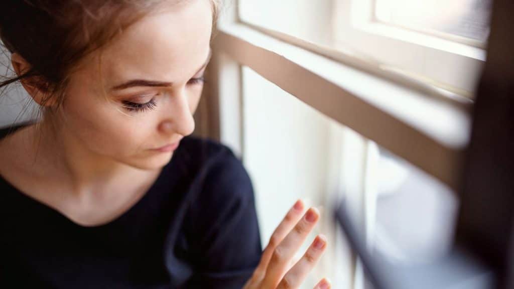 A young woman with a thoughtful expression looks down, her fingers resting on a windowsill.