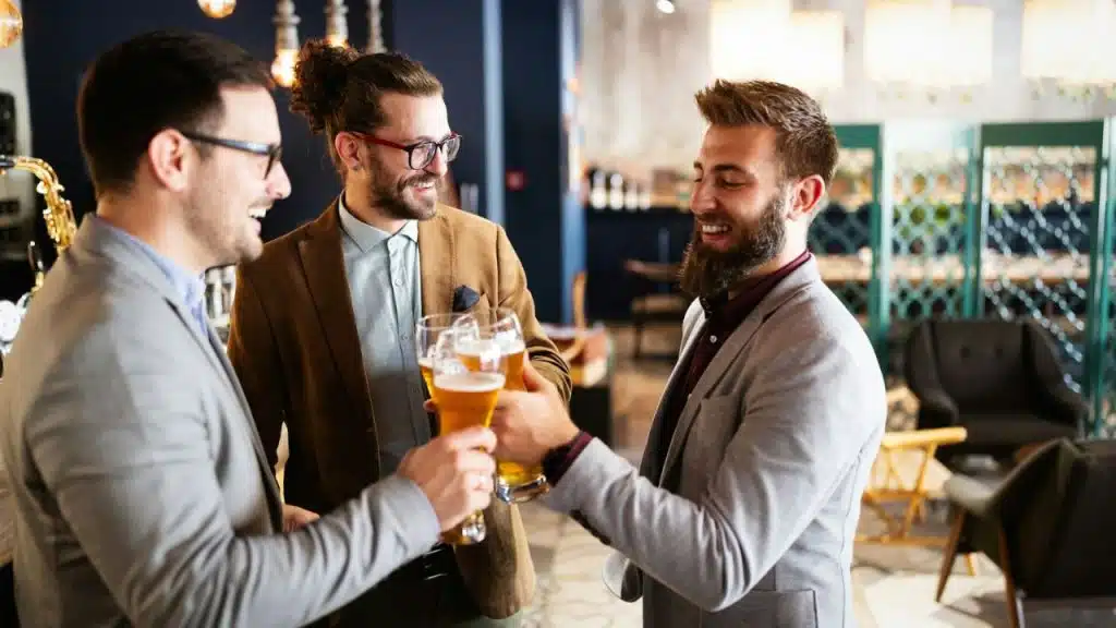 Three men in business casual attire cheerfully toasting with glasses of beer in a bar.