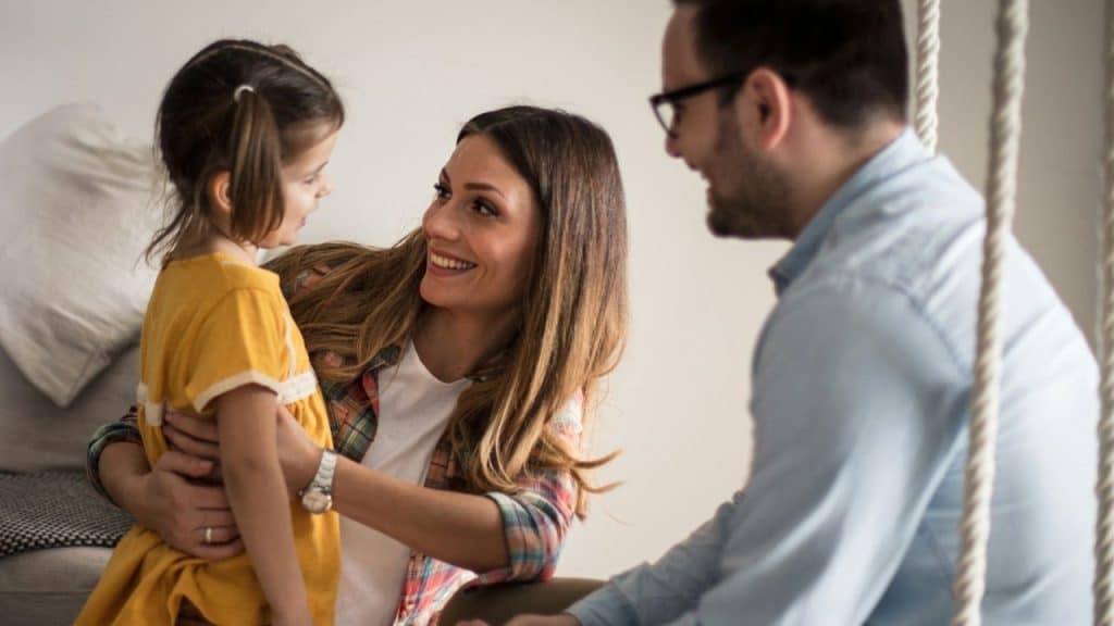 Smiling woman embracing a young girl in a yellow dress while a man watches and smiles.