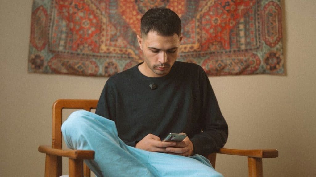 A man in a dark shirt and blue pants sits looking at his phone.