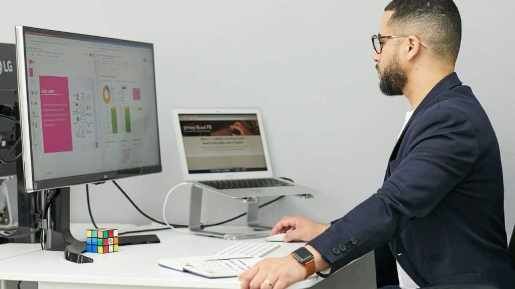 A man in a navy suit works at a desk with a monitor, laptop, and Rubik's cube.