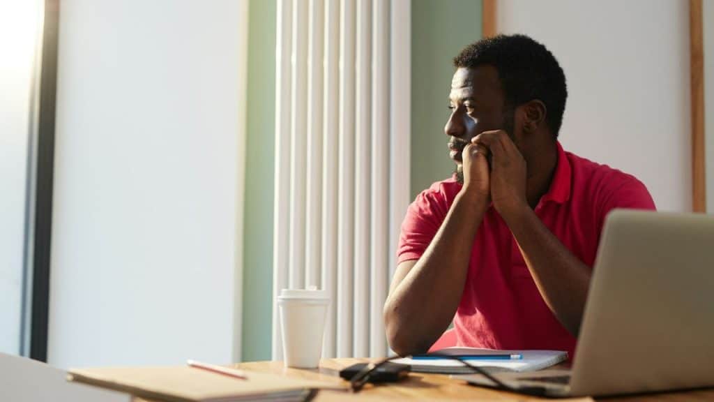 A Black man in a red shirt sits at a desk, looking out the window, his hands clasped.