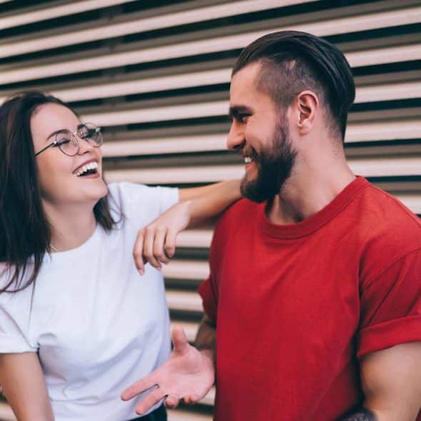 Smiling couple leaning against a slatted wall, the woman wearing glasses.