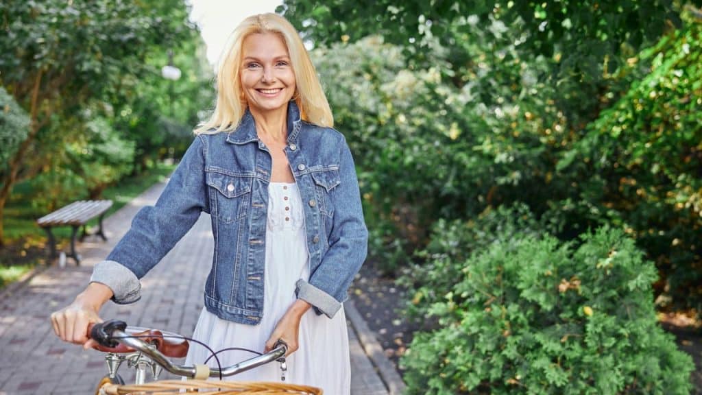 A smiling blonde woman stands on a path with a bicycle in a park.