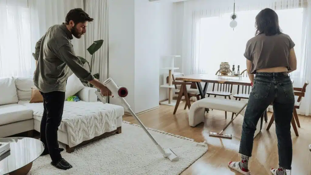 A man vacuums a shaggy rug while a woman stands nearby in a bright living room.
