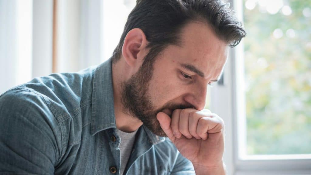 Close-up of a distressed man with a beard biting his fist near a window.