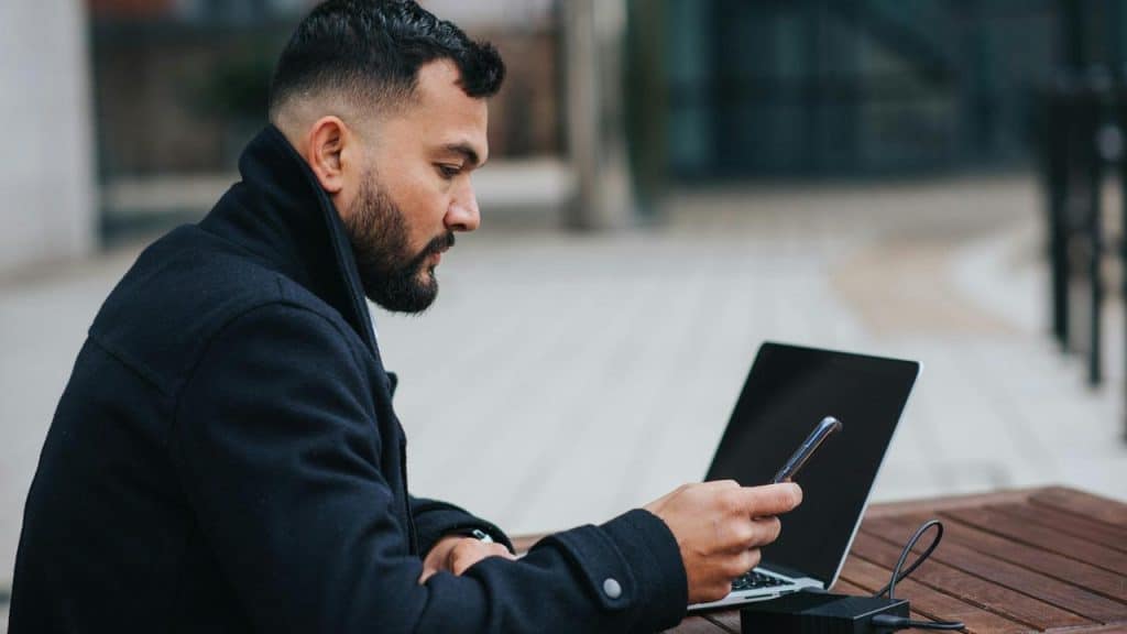 A man using a smartphone beside a laptop outdoors.